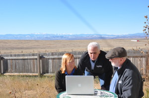 banner - Skyping in the Rockies - John Walczak, with 2 zoo workers, as they check laptop for images they are working on, background are beautiful snow capped mountains, with flat plains land behind the fence