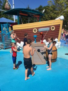 photo - group of boys and girls, in swim suits, playing with water spout element, at the splash park, the boat is in the background, on a sunny summer day