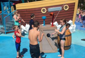 photo - group of boys and girls, in swim suits, playing with water spout element, at the splash park, the boat is in the background, on a sunny summer day