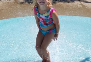 photo - little girl, in pretty bathing suit, smiling, playing in the zoo splash park on a sunny summer day