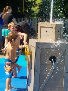 photo - couple of boys, in swim trunks, playing with water spout element in a box, while playing in splash park, on a sunny summer day