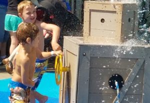 photo - couple of boys, in swim trunks, playing with water spout element in a box, while playing in splash park, on a sunny summer day