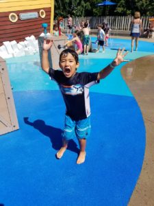photo - young man, in swim outfit, having fun, raised hands, yelling, while in splash park, background is more kids enjoying splash park too