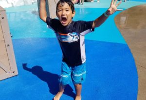 photo - young man, in swim outfit, having fun, raised hands, yelling, while in splash park, background is more kids enjoying splash park too