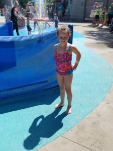 photo - young girl, in swim suit, standing inside splash park, with more kids, adults in the background, with water spouts going behind her