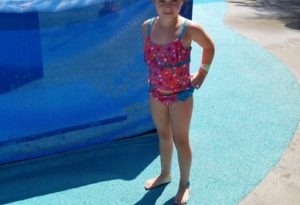 photo - young girl, in swim suit, standing inside splash park, with more kids, adults in the background, with water spouts going behind her