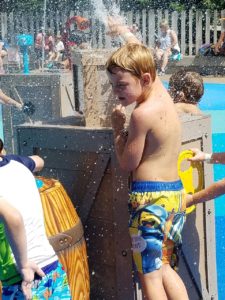 photo - young men, in swim trunks, playing with spouting water element, in splash park, with other children/adults in background