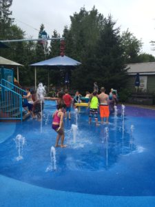 photo - children playing in underwater splash park, with individual water spouts coming from the ground, umbrellas, background of trees
