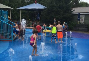 photo - children playing in underwater splash park, with individual water spouts coming from the ground, umbrellas, background of trees