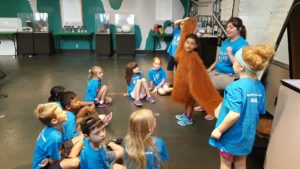 photo - boys and girls zoo day campers, with keeper counselor, displaying camper wearing costume of orangutan hair, which shows just how long their arms really are.