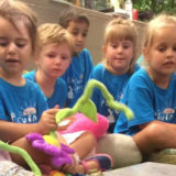 photo - group of pre k safari day campers, sitting, watching something going on in their circle, but some are holding plush flowers, frogs in the group