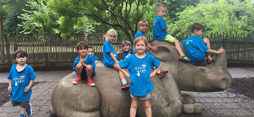 banner - boys and girls zoo day campers, in blue penguin cove t shirts, all sitting on rhino statue, all laughing, smiling
