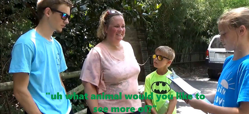 photo - zoo camper, asking a visitor their thoughts about what animal would they like to see more of, at the zoo, there is teenager and young child with the visitor,