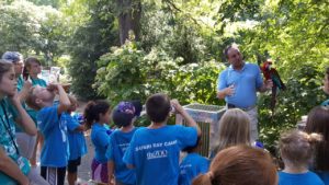 photo - bird keeper, showing a colorful red headed macaw, to a group of zoo day campers, wearing blue camper t shirts, on a sunny summer day, with counselors observing the group