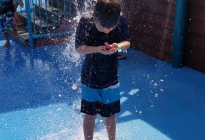 photo - young boy, in swimsuit, standing under waterfall at splash park in the zoo,