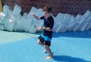 photo - young boy, in swimsuit, running thru water spray, at splash park at the zoo