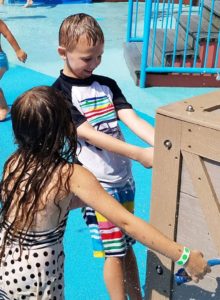 photo - young boy, girl playing with spout levers while in splash park, they are wearing colorful swimsuits