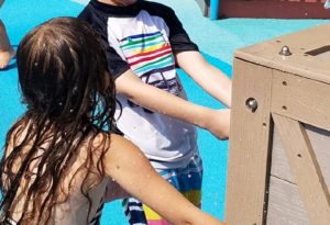photo - young boy, girl playing with spout levers while in splash park, they are wearing colorful swimsuits