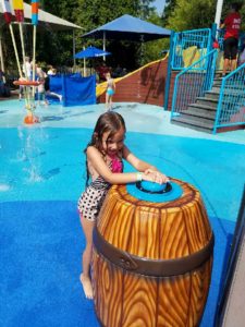 photo - young girl, in swimsuit, playing with brown barrel water spray spout, trying to keep water in, in zoo splash park, in background are more of water spouts, umbrellas for cover