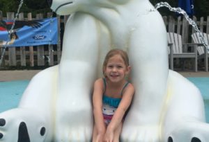 photo - young girl, sitting in arms of white sitting polar bear statue, in splash park water park, with water jumping around behind the bear
