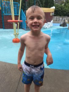 photo - little boy, in blue swim trunks, standing in splash park, smiling, having fun
