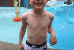 photo - little boy, in blue swim trunks, standing in splash park, smiling, having fun