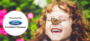 photo - blurred background of pink flowers and green bushes, with head shot of girl w/red hair, laughing, and a butterfly has landed on her nose, presented by Ford logo, Ford motor company highlighted in what circle