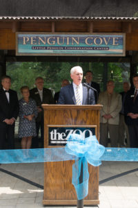 photo - john walczak, at podium, for penguin cove ribbon cutting, with audience of visitors behind him, on a bright sunny day