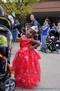 photo - little girl in red princess dress with designs, wearing bejeweled crown on head, standing by a baby stroller, with crowd of zoo visitors behind her on plaza