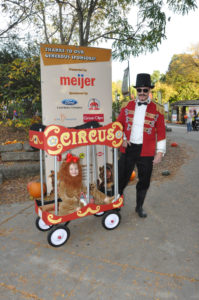 image - visitor dressed a circus ring leader, with wagon made into a lion cage, with small child, dressed as lion, sitting inside, with circus top display board, meijer bill board also in background