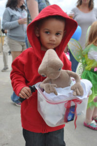 photo - young boy, dressed as Elliot from ET movie, in red hoodiie, holding handlebars with basket carrying brown ET, the little alien in the ET movie