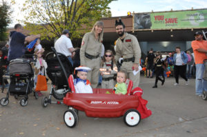 photo - 2 adults dressed in brown uniforms as ghost busters ; 1 child also dressed as a ghost buster; 1 child in wagon dressed as the marshmallow character in white uniform and sailor hat; 1 child sitting in the wagon dressed in green costume, on the plaza with lots of visitors in the background.