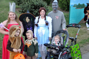 photo - family costumed as wizard of oz characters, dorothy; tin man; wicked witch of the west, good witch of the east, plus child dressed as the cowardly lion, child dressed as scarecrow, and child in stroller is a munchkin