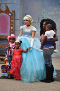 photo - cindrella, dressed in blue ball gown, with 2 children dressed as cowgirl in pink hat, neckerchief, and plaid pink shirt, another girl dressed in red princess dress, with bejeweled crown, with adult holding smaller child watching them all