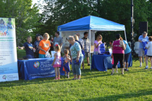 photo - display table for Norton's Childrens Hospital, and a blue tent, hospital employees giving out swag bags, info about norton's, to zoo guests inquiring about the info that is available, on lawn area, on a sunny summer day