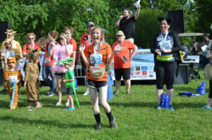 photo - young girl, with face paint tiger face, showing excitement, with other walkers, runners, visitors, some in costumes, also showing excitement, a male person talking on microphone, standing on a float for the race