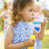 photo - blurred children background, with single little girl, standing in front, blowing bubbles, with 5-6 bubbles floating away, you can see the rainbow colors in the bubbles, on a sunny summer day