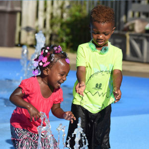 photo - 2 kids playing in splash park, with popup water spouts, smiling and laughing