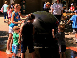photo - water ball on main plaza, with kids trying to stop it from rolling in the water, which you cannot stop, variety of visitors on the plaza.
