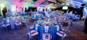 photo - inside oasis tent, setup for private event with variety of tables, chairs, decorated with blue, white table clothes, chair covers, dishes of food on table, ceiling has multi colors, visitors can be seen throughout the location