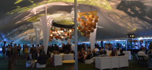 photo - oasis tent holding a night time event with multi lighting designs in ceiling, white chairs and couches with tables for drinks, food, variety of guests milling around or sitting throughout the tent