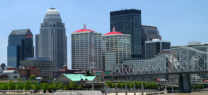 banner - downtown louisvillel, galt house hotels w/red roofs, humana building, variety of other tall buildings, 2nd street bridge, and joe's crab shack w/green roof, at the downtown waterfront, lined with blooming green trees