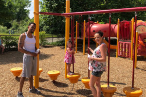 photo - mom, dad and child at playground, child playing on step/pole equipment, everyone is smiling, on a sunny summer day