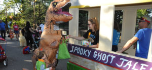photo - trick or treat booth, volunteers giving out candy, to costumed zoo visitors, a dinosaur, and other visitors attending halloween at the zoo