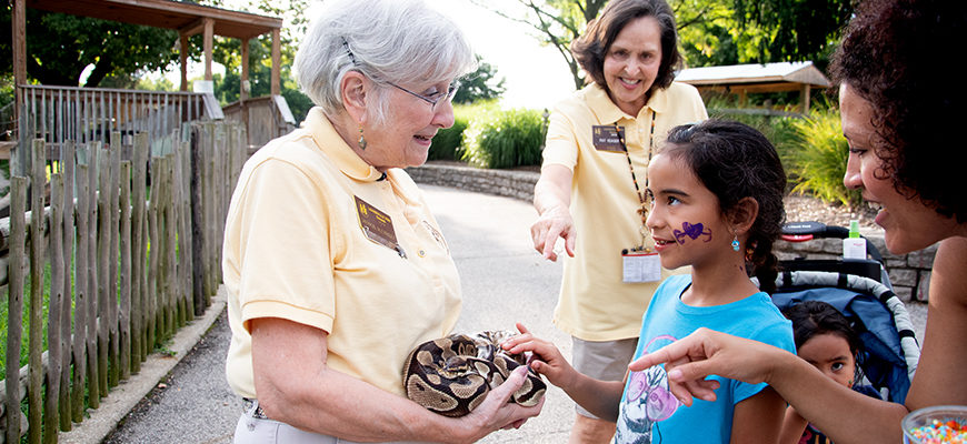 photo - keeper showing a brown w/individual size brown markings on its body, with another keeper telling the 3 visitors, one of which is child petting the snake, info about the snake, background is pathway thru the zoo