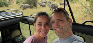 photo - Fred Hougland, and friend, smiling, while watching herd of elephants in background from their observation vehicle in Tanzania