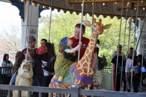 photo - shows couple of dads, helping their kids , on carousel ride, one is riding on a lynx, other is on a giraffe, background shows other visitors standing around carousel, or on the carousel