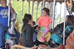 photo - female standing next to child, who is riding a carousel black horse, adult male in background riding a horse