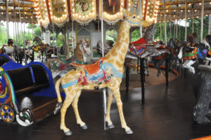 photo - carousel animal giraffe, tan with yellow markings, orange saddle, background shows variety of other carousel animals that you can ride