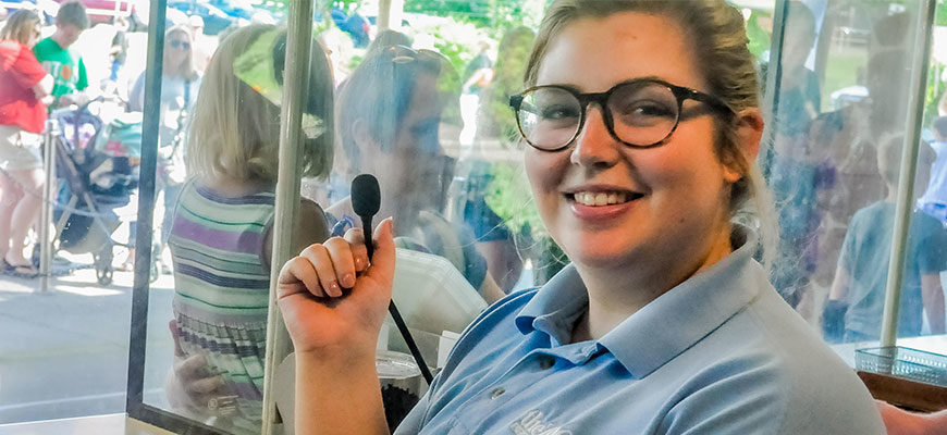 image - Zoo employee, working admission windows, wearing blue zoo work short, nice smile, holding microphone, and background is view of visitors at the windows buying tickets.